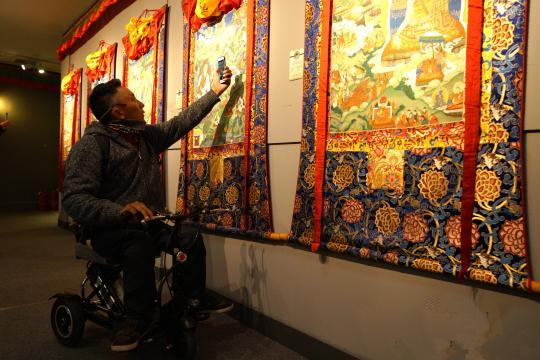 西藏安多强巴唐卡艺术画院成立一周年师徒精品作品展在拉萨举行。图为11日，一位腿部残疾的唐卡画师参观展览并用手机记录写实派唐卡特点。