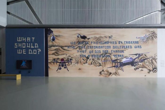 新委任创作，Chto Delat，枯石与相机的寓言，墙上水墨，6×16 m，2018，致谢：PSA，画面译文:相机拍下了我们 / 向我们传达出一种讯息 / 千年来我们都未曾改变